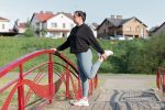 A woman in active wear performs a stretching exercise on a red bridge in a suburban park setting.