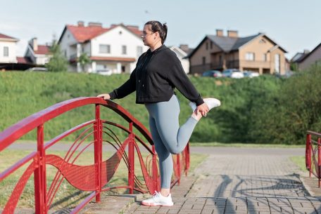 A woman in active wear performs a stretching exercise on a red bridge in a suburban park setting.